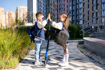Happy sibling children with school backpack stand with scooter and give high five in front of multi-storey building city background. Eco-friendly urban transport for kid. High quality