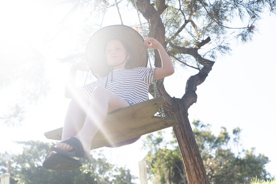 Girl swinging on a handmade swing wearing a big broad brim hat in summer sunshine