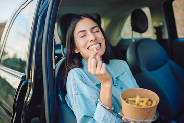 Happy female driver eating french fries from a takeaway box inside a car, enjoying a tasty snack on the go