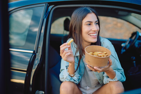 Happy young woman enjoying some french fries from a takeaway box while sitting in her car, smiling and looking away
