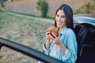 Happy young woman enjoying a burger during a break from driving, leaning out of her car window in a...