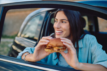 Young woman enjoying a delicious hamburger while sitting in her car, taking a break during an adventurous road trip