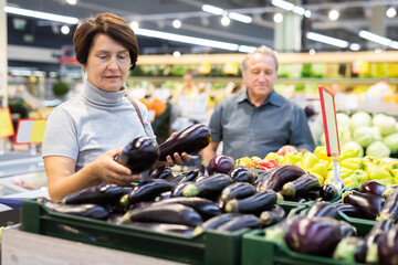Elderly buyer woman chooses ripe eggplant on grocery store window