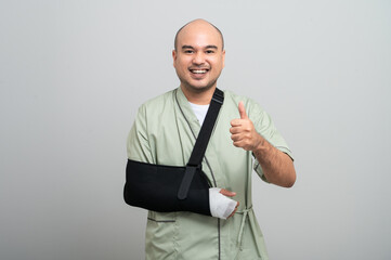 Asian man in patient attire smiles gently at the camera, wearing an arm sling and bandage, striking...