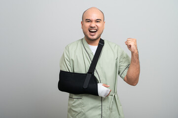 Asian man in patient attire smiles gently at the camera, wearing an arm sling and bandage, striking...