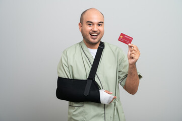 An Asian man in a hospital gown smiles gently at the camera with his arm bandaged and bandaged,...