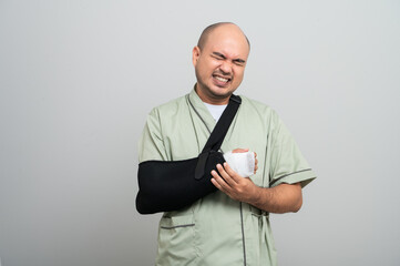 Asian man in patient attire smiles gently at the camera, wearing an arm sling and bandage, striking...