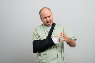Asian man in patient attire smiles gently at the camera, wearing an arm sling and bandage, striking...