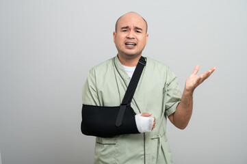 Asian man in patient attire smiles gently at the camera, wearing an arm sling and bandage, striking...