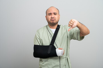 Asian man in patient attire smiles gently at the camera, wearing an arm sling and bandage, striking...
