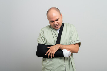 Asian man in patient attire smiles gently at the camera, wearing an arm sling and bandage, striking...