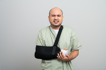 Asian man in patient attire smiles gently at the camera, wearing an arm sling and bandage, striking different poses to show resilience and a positive attitude during recovery.