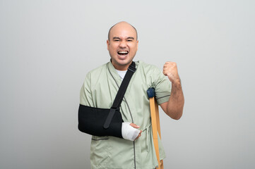 Asian man in hospital gown smiles gently at camera with arm bandages and bandages and crutches, posing to show resilience and positive attitude during recovery.