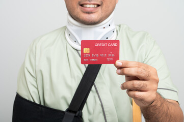 Asian man in hospital gown smiles gently at camera with arm bandages and bandages and crutches, posing holding health insurance card to demonstrate resilience and positive attitude during recovery.
