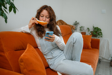 Young woman enjoys a slice of fresh melon while browsing on her smartphone, relaxing on a comfortable orange sofa in her cozy living room, with her ginger cat resting nearby