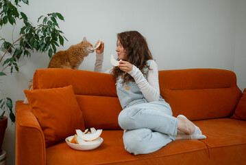 Young woman sitting comfortably on an orange sofa, sharing juicy slices of fresh melon with her playful ginger cat in a warm and inviting home environment