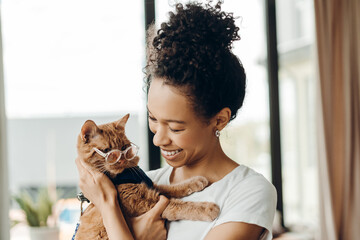 Portrait of young caring African American woman hugging ginger cat with harness and glasses at home