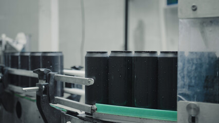 Black aluminum cans with water drops are moving on a conveyor belt inside a beverage factory, suggesting an industrial process of bottling or packaging drinks