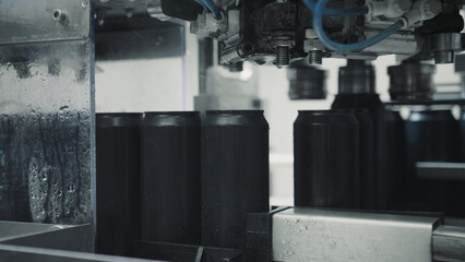 Black aluminum cans are moving along a conveyor belt in a beverage factory, undergoing a filling and sealing process under the watchful eye of automated machinery