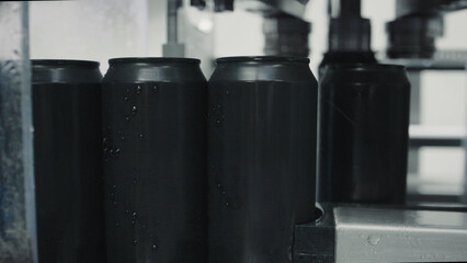 Black aluminum beverage cans with condensation droplets are moving along a conveyor belt inside a...
