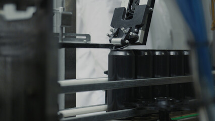 Automated machinery placing lids onto a row of black aluminum cans, moving along a conveyor belt in a beverage production facility, demonstrating modern industrial automation