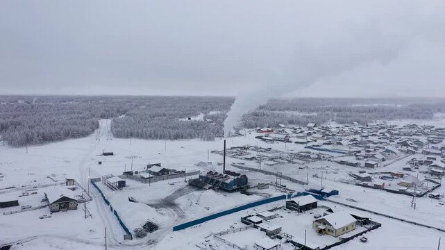 Aerial view of snow-laden buildings with smoke rising from a chimney, creating a stark contrast against the white landscape, Oymyakon, Sakha Republic, Russia.