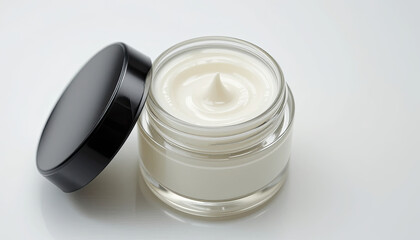 Close up of a glass jar filled with white cream and a black lid resting beside it on a white surface