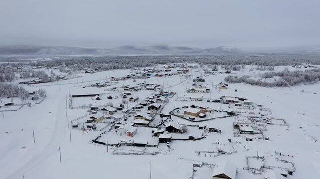 Aerial view of a small settlement's buildings, all blanketed in thick snow, with a forest and distant mountains under a cloudy sky, Oymyakon, Sakha Republic, Russia.