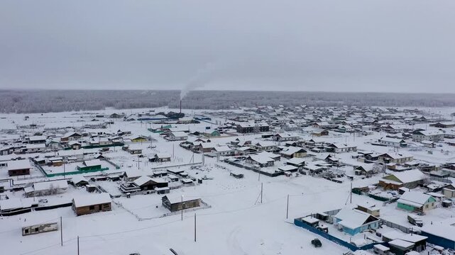 Aerial view of snow-covered houses and a factory releasing smoke into the sky, creating a stark contrast against the white landscape, Oymyakon, Sakha Republic, Russia.