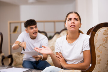 Angry young husband, holding documents in his hands, swears at his wife in the apartment and reproaches her for inattention