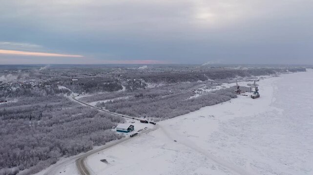 Aerial view of snow-covered trees and a frozen river with cranes, creating a stark contrast against the muted sky, Yakutsk, Sakha Republic, Russia.