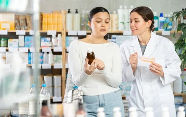 Gordijnen Asian woman buyer choosing medicinal cough liquid syrup standing between shelves at big pharmacy, young girl pharmacist consulting customer © JackF