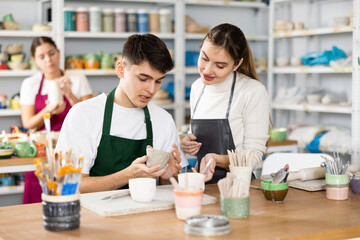 Interested young man and woman in aprons making handmade cups of row clay material and master...