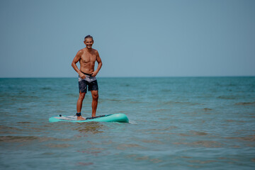 A young Caucasian man with short brown hair stands on a paddleboard in the ocean. He wears black...