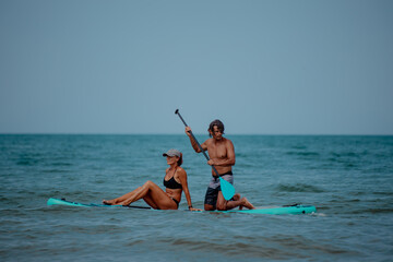 A young Caucasian man and a young Hispanic woman enjoy paddleboarding in the ocean. The man holds a...