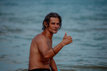A middle-aged Caucasian man with curly hair gives a thumbs-up while standing in the ocean. The water is calm and blue, creating a relaxed beach atmosphere.