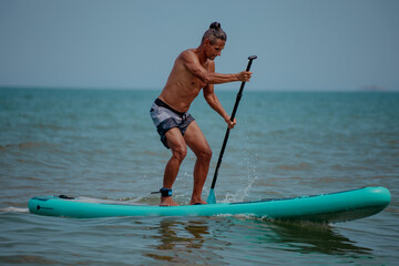 A young Caucasian man with short brown hair stands on a paddleboard in the ocean. He wears black...