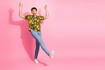Cheerful young man in tropical shirt dancing against pink background with headphones enjoying...