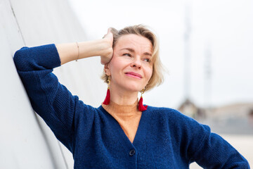 woman with short blonde hair and red tassel earrings leans against a wall, looking off to the side...