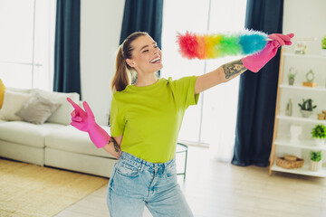Enthusiastic young woman cleaning her modern living room with a rainbow feather duster and wearing...