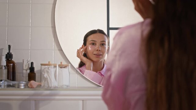 Reflection in mirror of young beautiful brunette woman putting under eye patches, looking at herself, touching face and smiling during self care ritual