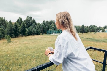 female silhouette on balcony with coffee cup, back view against scenic rural field and overcast sky, concept of mindfulness, nature connection, seasonal getaway