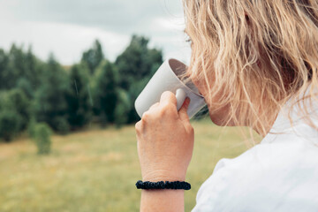close up of woman drinking from white cup with blurred green forest in background, concept of slow living, rainy morning, rural wellness