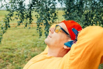 man in yellow hoodie and red cap resting with closed eyes on wooden bench under birch trees during warm summer day in rural outdoor area, vacation travel wellness retreat