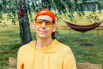 man looking at camera in casual outfit, orange-red color palette, sunglasses reflecting light, outdoor greenery and trees, concept of youth expression, streetwear blog, nature leisure