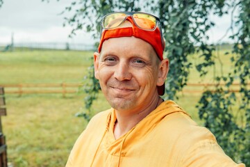 smiling man in orange clothes, cap, looking at camera, bright colors and relaxed outdoor setting with trees and hammock in background, casual fashion, men s accessories, summer style