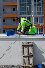 Fototapeta premium Construction Worker Performing Maintenance on Rooftop While Wearing Safety Gear in Urban Setting with Modern Apartments in Background Under Clear Blue Sky