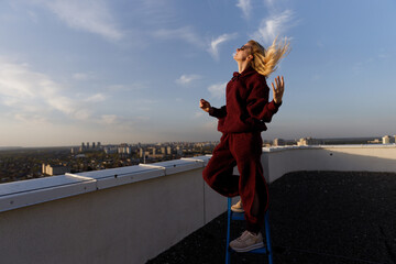 A Young Woman Standing on a Chair on a Rooftop Enjoys the Breeze in a Cozy Outfit While Overlooking...