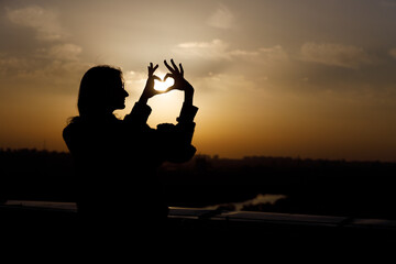 Silhouette of a Woman Making a Heart Gesture Against a Golden Sunset Sky with Cloud Patterns...