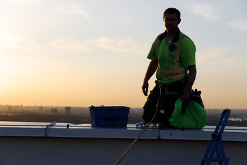 A Rooftop Worker in a Safety Harness at Dusk, Showcasing the Courageous and Daring Work of...